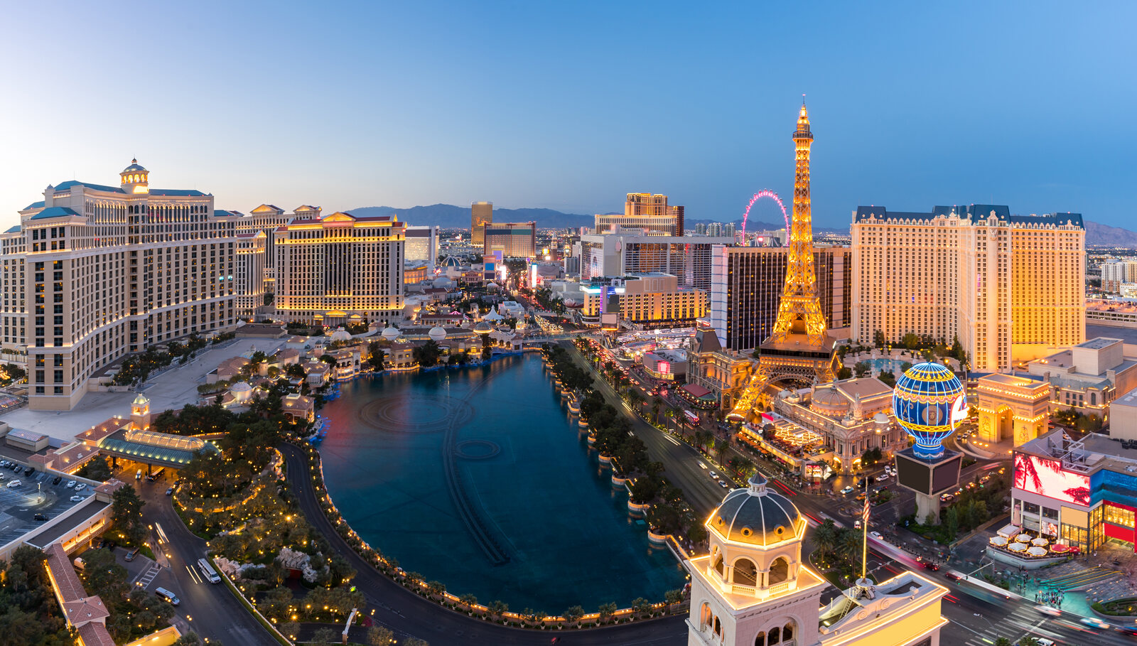 Las Vegas Strip during twilight, featuring the illuminated Eiffel Tower replica at Paris Las Vegas, the vibrant High Roller observation wheel, and the dancing fountains of the Bellagio Hotel. | Houfy — No Fees, Book Direct