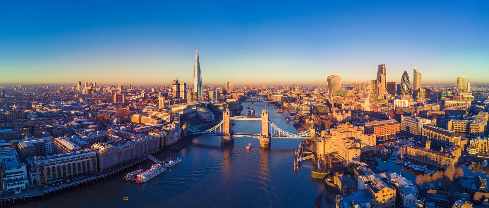 London skyline at sunrise highlights the iconic Tower Bridge over the River Thames, with The Shard and modern skyscrapers in the background, bathed in warm, golden sunlight. | Houfy — No Fees, Book Direct
