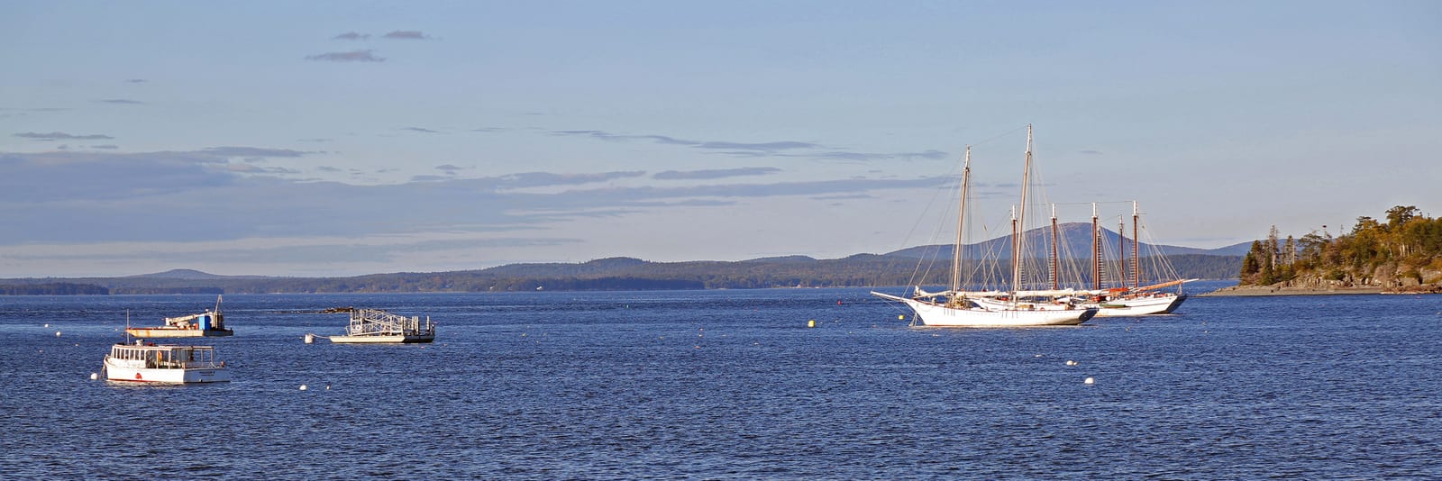 Sailboats gently float on the waters of Bar Harbor, Maine, with a backdrop of rolling hills and a clear sky, highlighting the tranquil charm and natural beauty of this picturesque coastal destination. | Houfy — No Fees, Book Direct