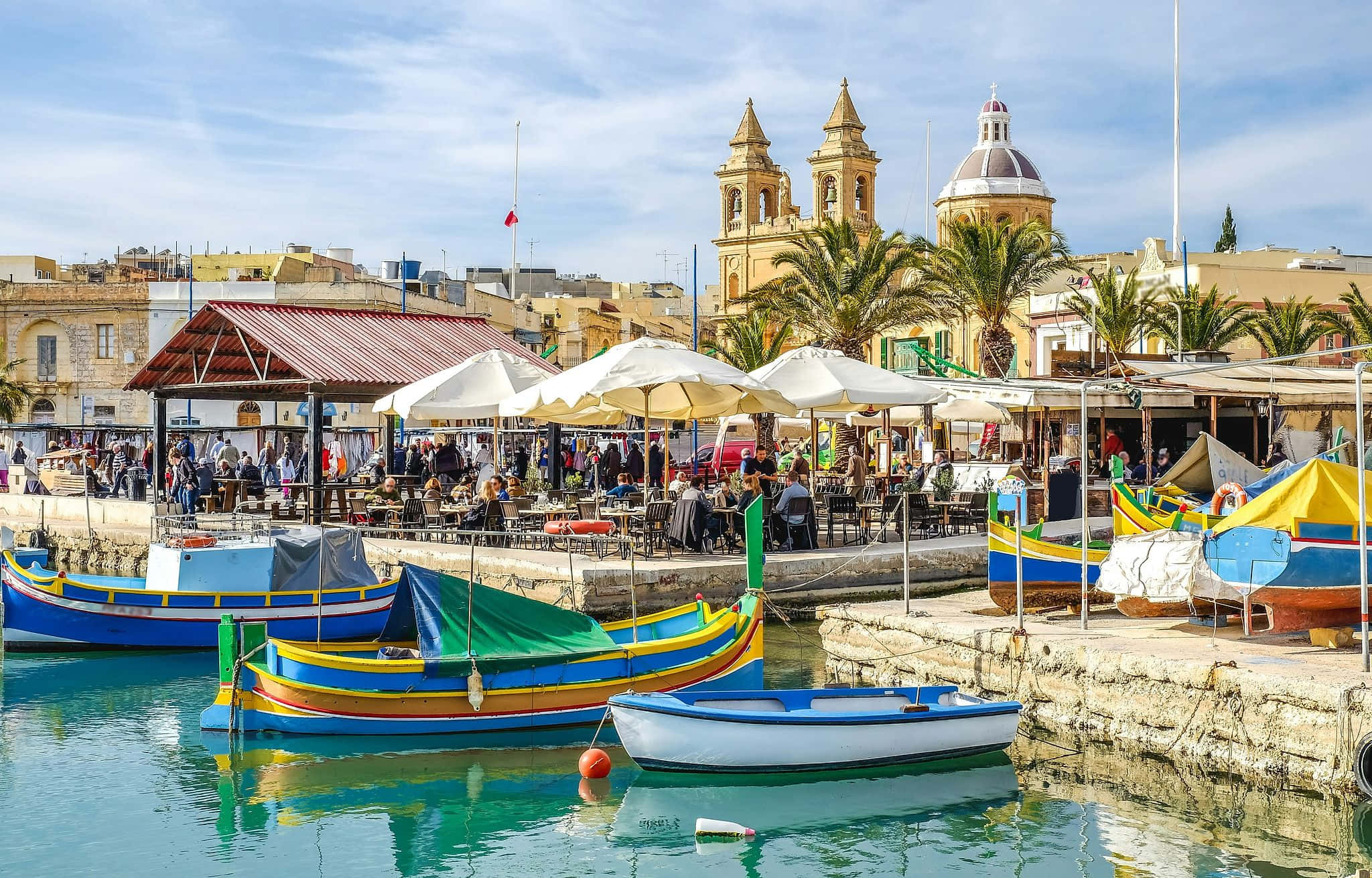 Marsaxlokk harbor, Malta, lined with vivid traditional Luzzu fishing boats in bright blue, yellow, and red with the Eye of Osiris painted on their bows, calm turquoise Mediterranean waters, outdoor cafe umbrellas along the stone promenade, and the golden honey-limestone towers and dome of the Marsaxlokk Parish Church under a clear pale blue sky — book a fee-free Malta vacation rental on Houfy | Houfy — No Fees, Book Direct