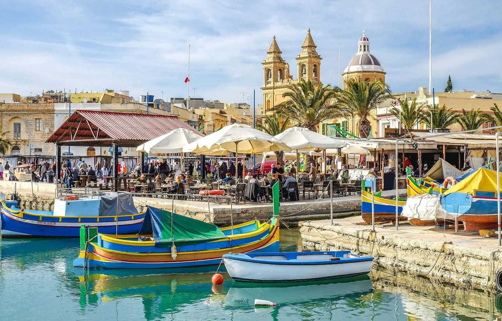 Marsaxlokk harbor, Malta, lined with vivid traditional Luzzu fishing boats in bright blue, yellow, and red with the Eye of Osiris painted on their bows, calm turquoise Mediterranean waters, outdoor cafe umbrellas along the stone promenade, and the golden honey-limestone towers and dome of the Marsaxlokk Parish Church under a clear pale blue sky — book a fee-free Malta vacation rental on Houfy | Houfy — No Fees, Book Direct