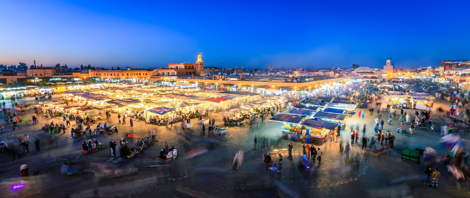 Vibrant evening scene of Jemaa el-Fnaa square in Marrakech, Morocco, bustling with market stalls and visitors under a clear, deep blue sky, capturing the lively atmosphere of this iconic destination. | Houfy — No Fees, Book Direct