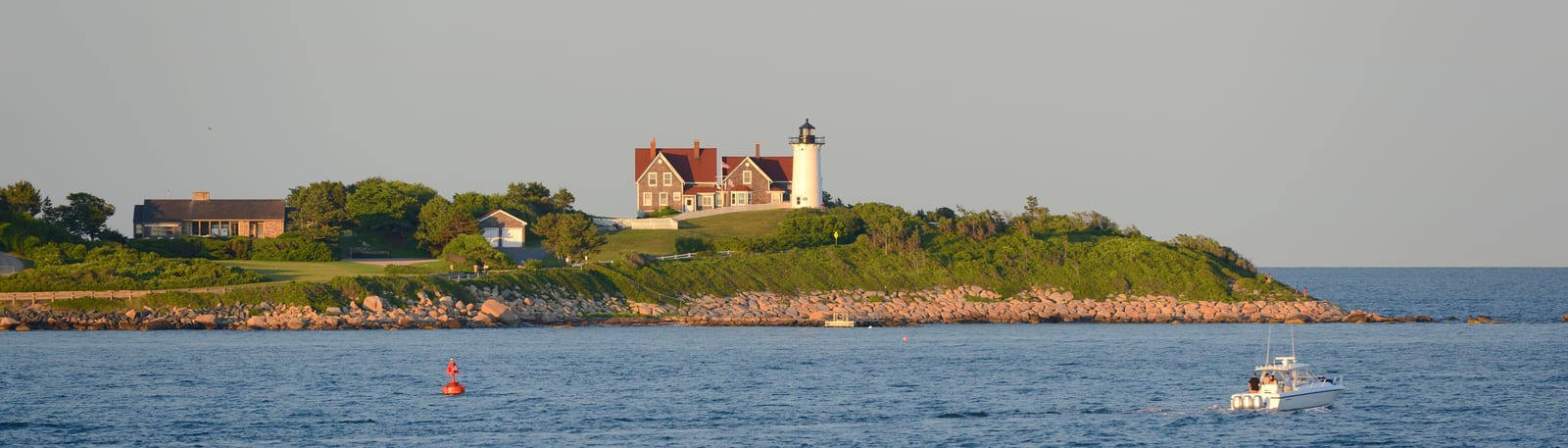 Nubble Lighthouse stands majestically on a rocky promontory surrounded by lush greenery and the Atlantic Ocean, with a charming boat navigating the waters near York, Maine, under the warm golden light. | Houfy — No Fees, Book Direct