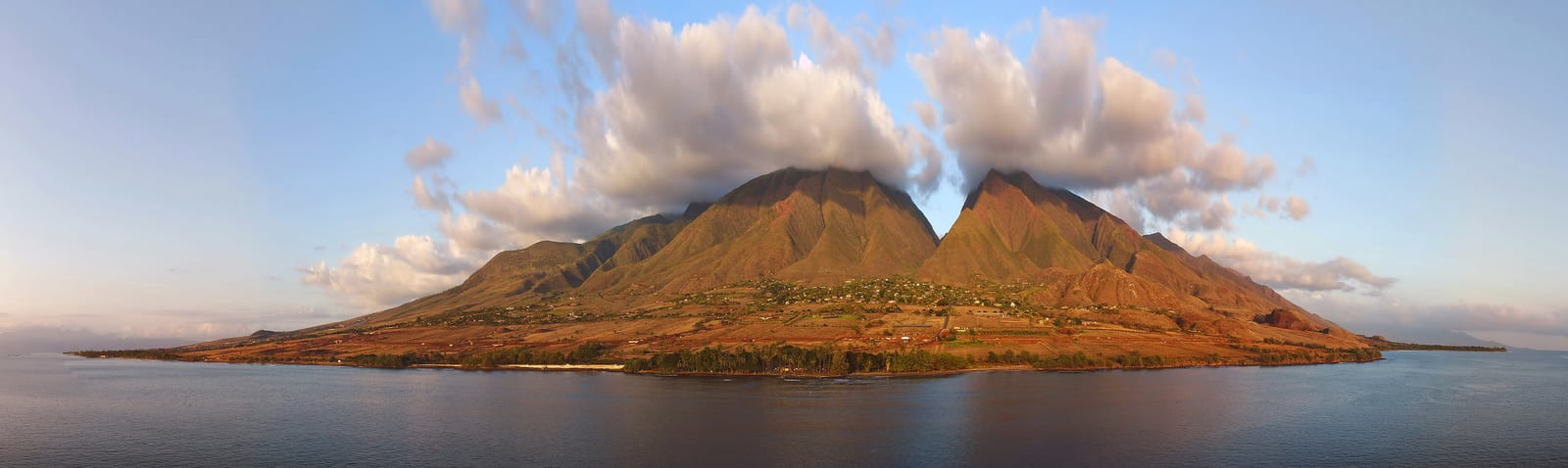 Maui's coastal panorama reveals twin volcanic peaks shrouded in clouds, contrasting with lush greenery and clear waters, capturing Hawaii’s unique island allure. | Houfy — No Fees, Book Direct