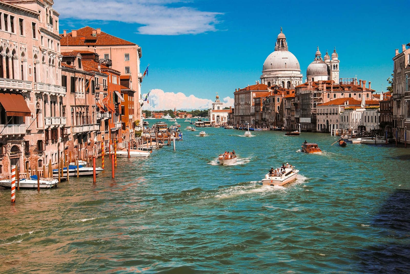 High-angle view of Venice's Grand Canal lined with terracotta and cream-colored palazzos, striped mooring poles, bustling water taxis and gondolas on turquoise water, and the iconic white Baroque domes of Santa Maria della Salute under a brilliant blue sky — book a fee-free Venice vacation rental on Houfy | Houfy — No Fees, Book Direct
