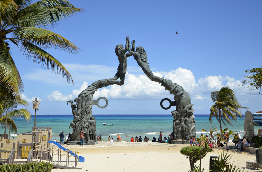 Beachfront sculpture featuring two figures reaching towards each other, forming an arch in Playa del Carmen, with palm trees and turquoise ocean creating a picturesque backdrop.