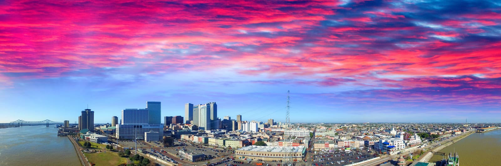Vibrant pink and blue sky over New Orleans skyline, featuring tall modern buildings, historic neighborhoods, and the Mississippi River with the iconic Crescent City Connection Bridge. | Houfy — No Fees, Book Direct