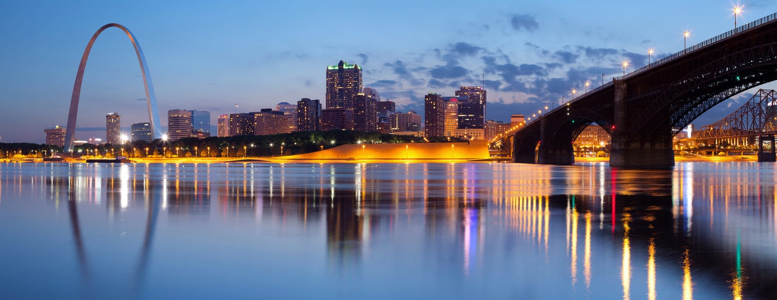 St. Louis skyline with the iconic Gateway Arch, illuminated against a twilight sky, reflecting on the calm Mississippi River, alongside the illuminated arches of the historic Eads Bridge. | Houfy — No Fees, Book Direct