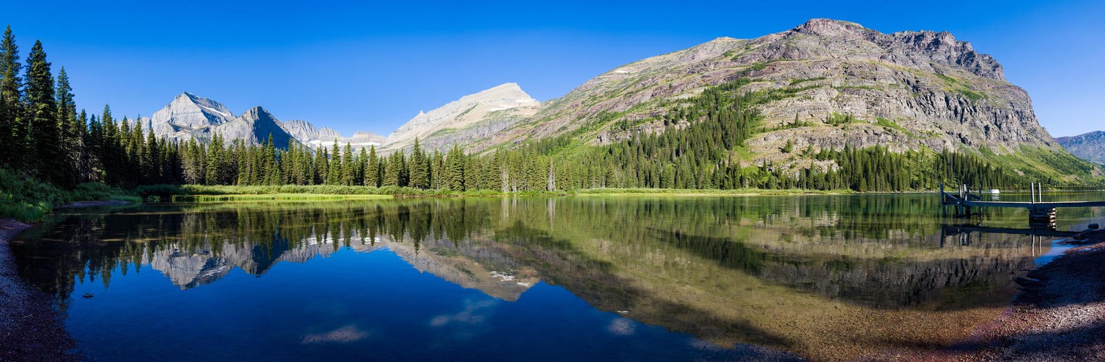 Pristine mountain range reflection in clear waters at Glacier National Park, framed by lush pine trees, with a wooden dock extending into the tranquil lake beneath a cloudless sky. | Houfy — No Fees, Book Direct