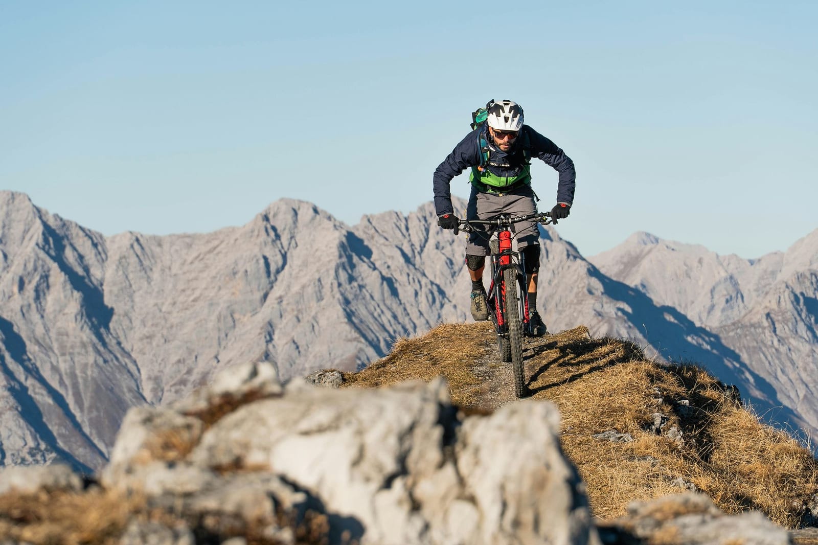 Male mountain biker on a red full-suspension bike navigating a narrow single track ridge trail at golden hour, with jagged blue-grey alpine peaks receding into the distance under a clear sky — book a fee-free mountain biking vacation rental on Houfy | Houfy — No Fees, Book Direct