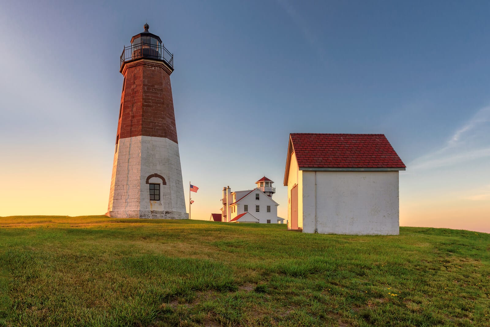 Lighthouse with red brick and white base stands tall beside a historic seaside building and a small shed at sunset in Narragansett, Rhode Island, with vibrant skies creating an inviting coastal scene. | Houfy — No Fees, Book Direct