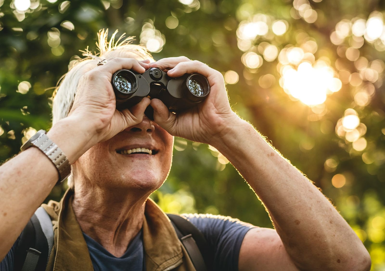 A joyful senior man in a tan outdoor vest smiling while scanning a sun-dappled forest canopy through binoculars at golden hour, with warm backlit foliage and bokeh light spots — book a fee-free nature viewing vacation rental on Houfy | Houfy — No Fees, Book Direct
