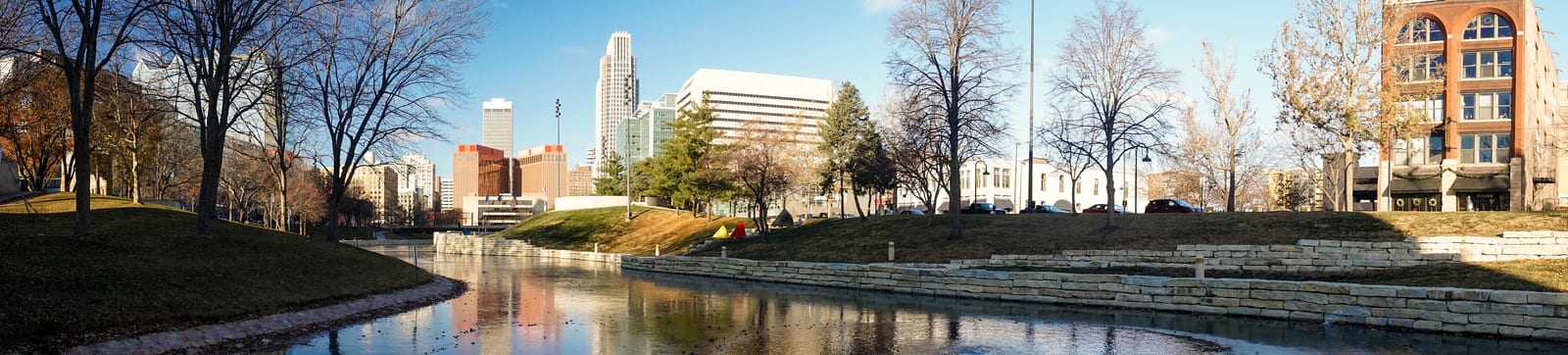 Downtown Omaha skyline with modern buildings standing tall against a clear blue sky, reflected in a serene waterway flanked by stone walls and leafless winter trees. | Houfy — No Fees, Book Direct