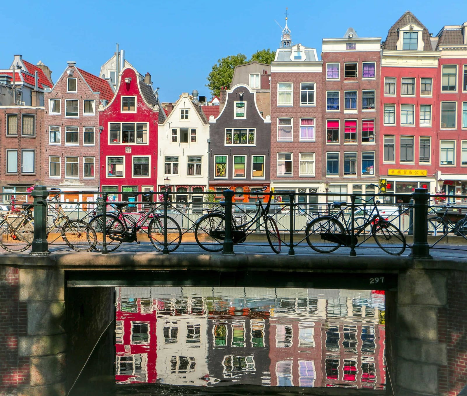 Colorful Amsterdam canal houses with stepped, neck, and bell gables in crimson, white, and brick reflected in still canal water beneath a stone bridge numbered 297, with a row of bicycles parked on the iron railing under a clear blue sky — book a fee-free Netherlands vacation rental on Houfy | Houfy — No Fees, Book Direct
