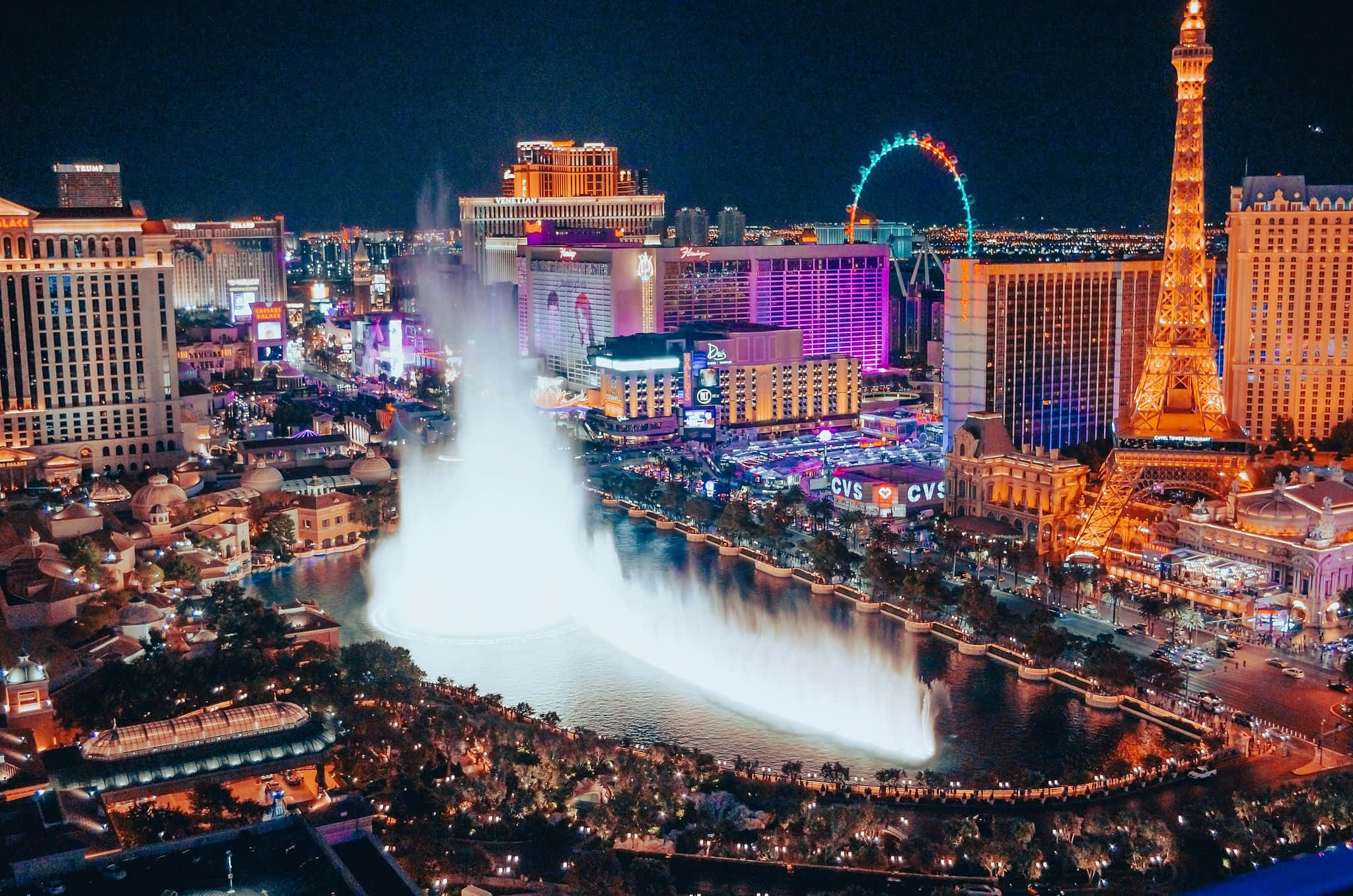 Aerial nighttime view of the Las Vegas Strip, Nevada, with the Bellagio Fountains in full spray, the glowing Paris Las Vegas Eiffel Tower, and the neon-lit High Roller observation wheel under a black sky — book a fee-free Nevada vacation rental on Houfy | Houfy — No Fees, Book Direct