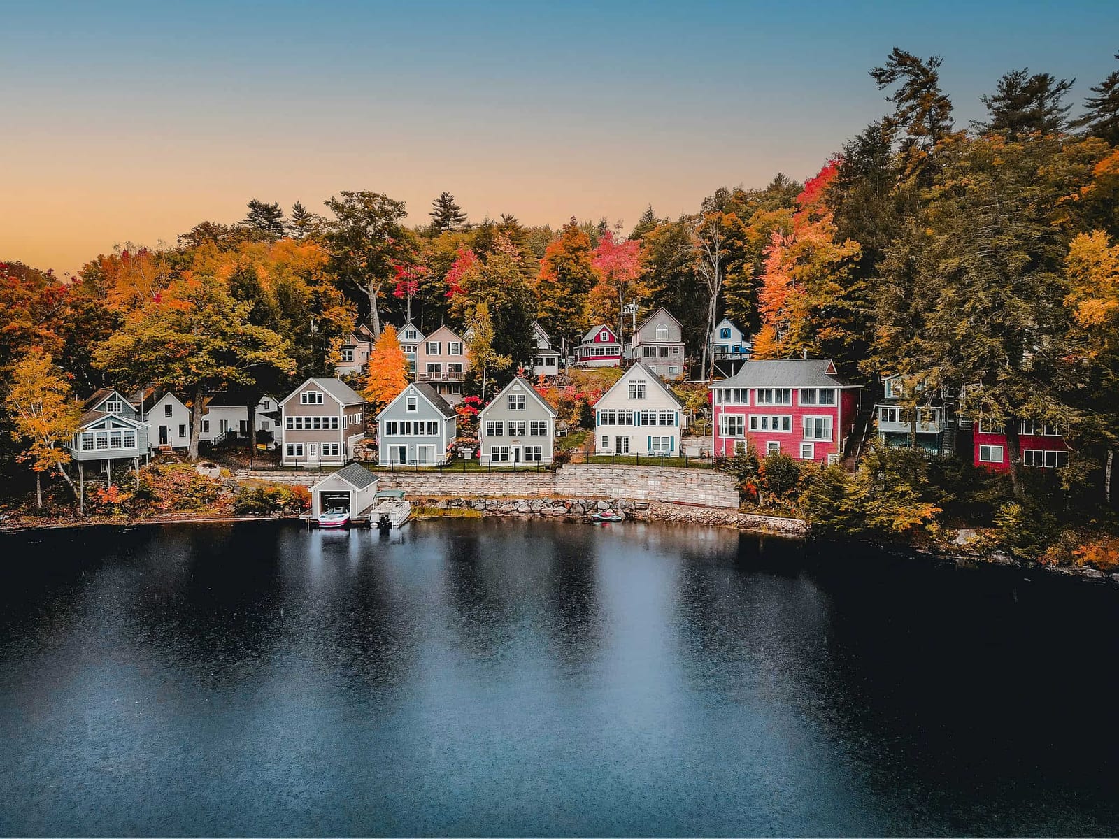 Row of classic New England lakefront cottages in New Hampshire's Lakes Region at golden hour during peak fall foliage, with white, seafoam green, pale blue, and barn-red Craftsman homes lining a stone seawall, a white boathouse at the water's edge, and a steep hillside blazing in fiery red, burnt orange, and gold reflected in the dark still lake water — book a fee-free New Hampshire vacation rental on Houfy | Houfy — No Fees, Book Direct