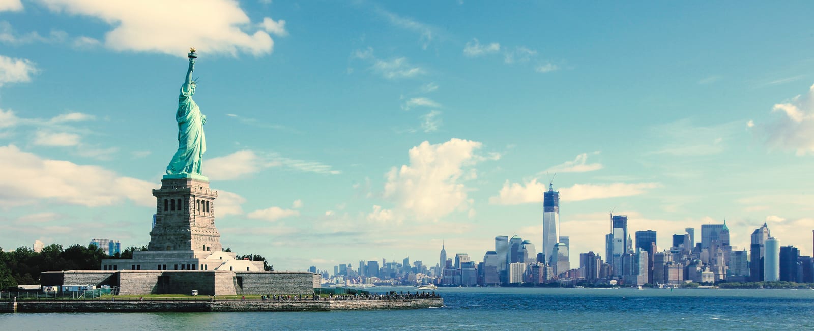 The Statue of Liberty stands tall beside New York City's iconic skyline under a bright blue sky, with the tranquil waters of the Hudson River enhancing the city's majestic allure. | Houfy — No Fees, Book Direct