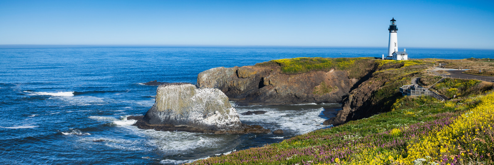 Yaquina Head Lighthouse stands majestically atop rugged cliffs overlooking the Pacific Ocean in Oregon, surrounded by vibrant wildflowers and rocky coastal formations. | Houfy — No Fees, Book Direct