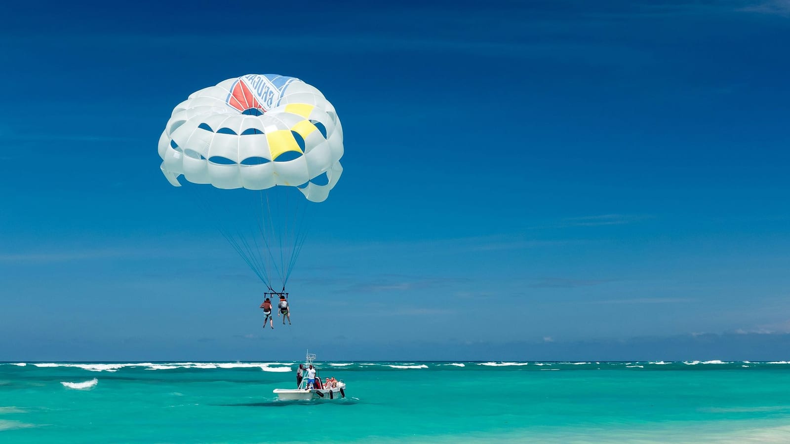 Two people tandem parasailing high above brilliant turquoise and aquamarine tropical ocean waters, suspended from a white dome parachute towed by a motorboat under a vast clear blue sky — book a fee-free parasailing vacation rental on Houfy | Houfy — No Fees, Book Direct