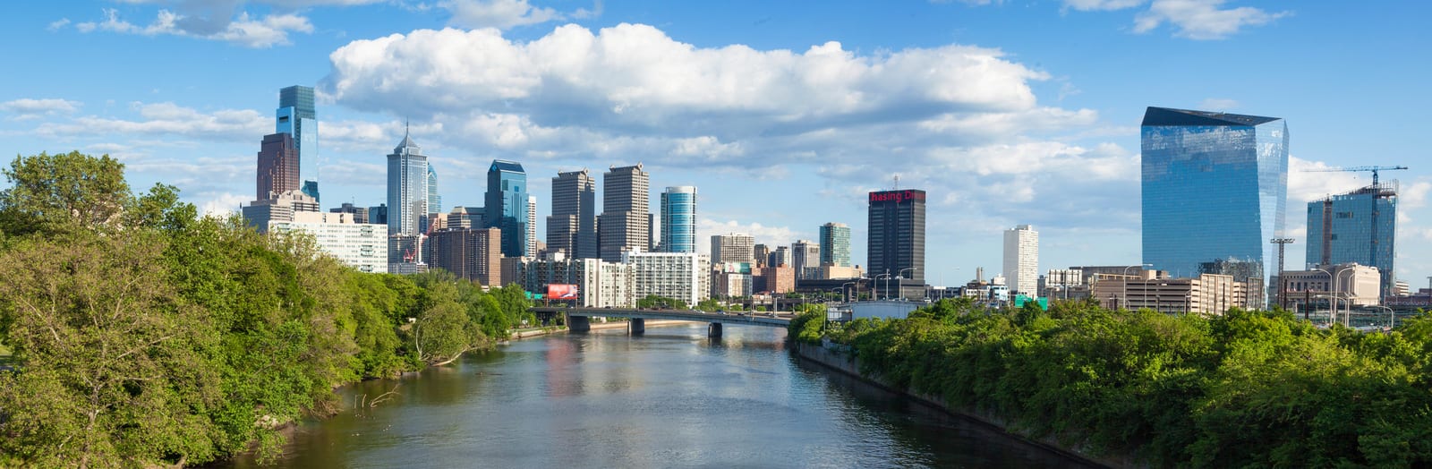 Philadelphia skyline view with skyscrapers and modern buildings reflected in the Schuylkill River, framed by lush green trees and a bridge, under a vibrant blue sky with fluffy clouds. | Houfy — No Fees, Book Direct