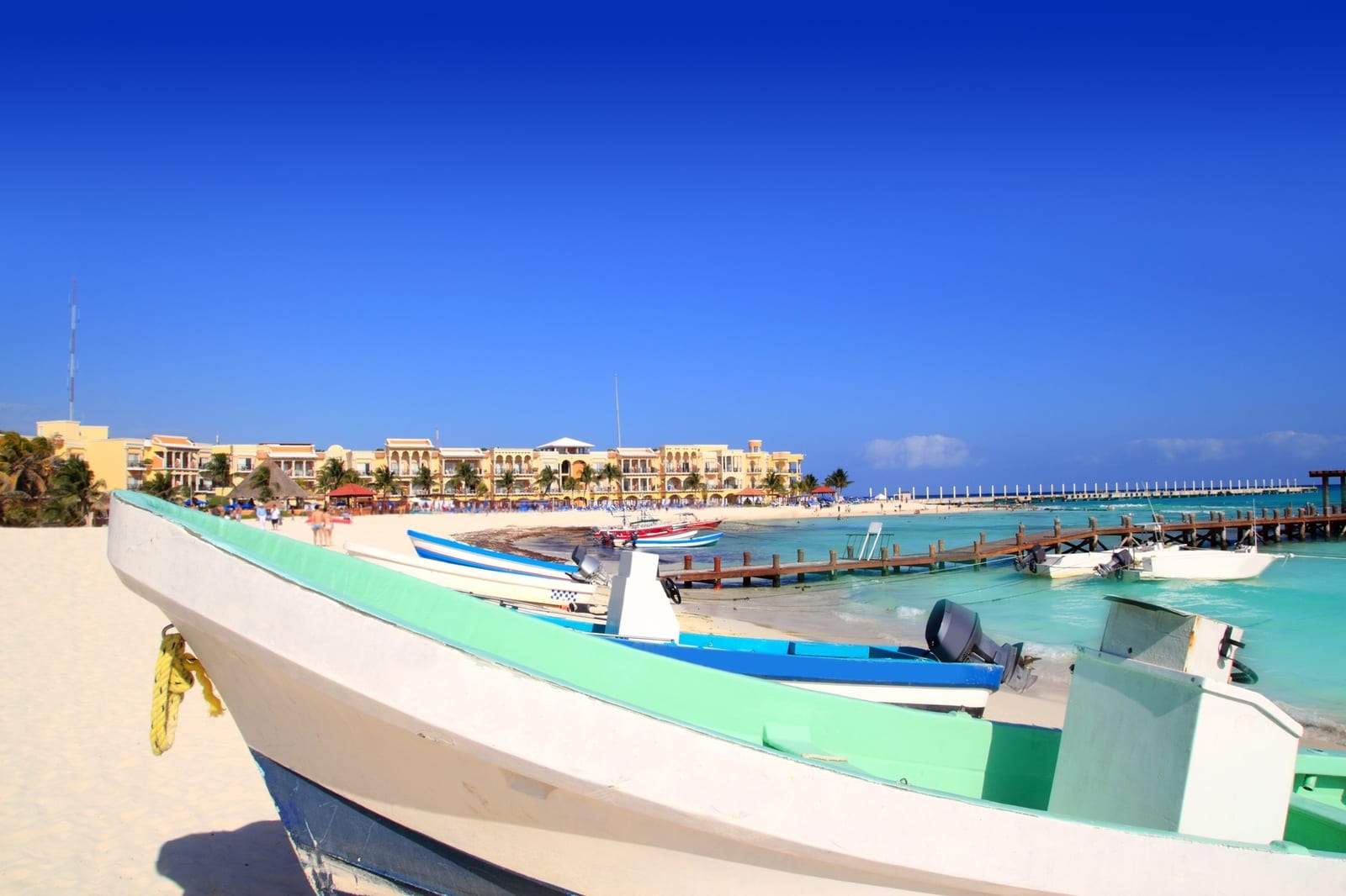 Pristine Playa del Carmen beach features colorful fishing boats on white sand against a resort backdrop, with a wooden pier stretching into turquoise Caribbean waters and clear blue skies. | Houfy — No Fees, Book Direct