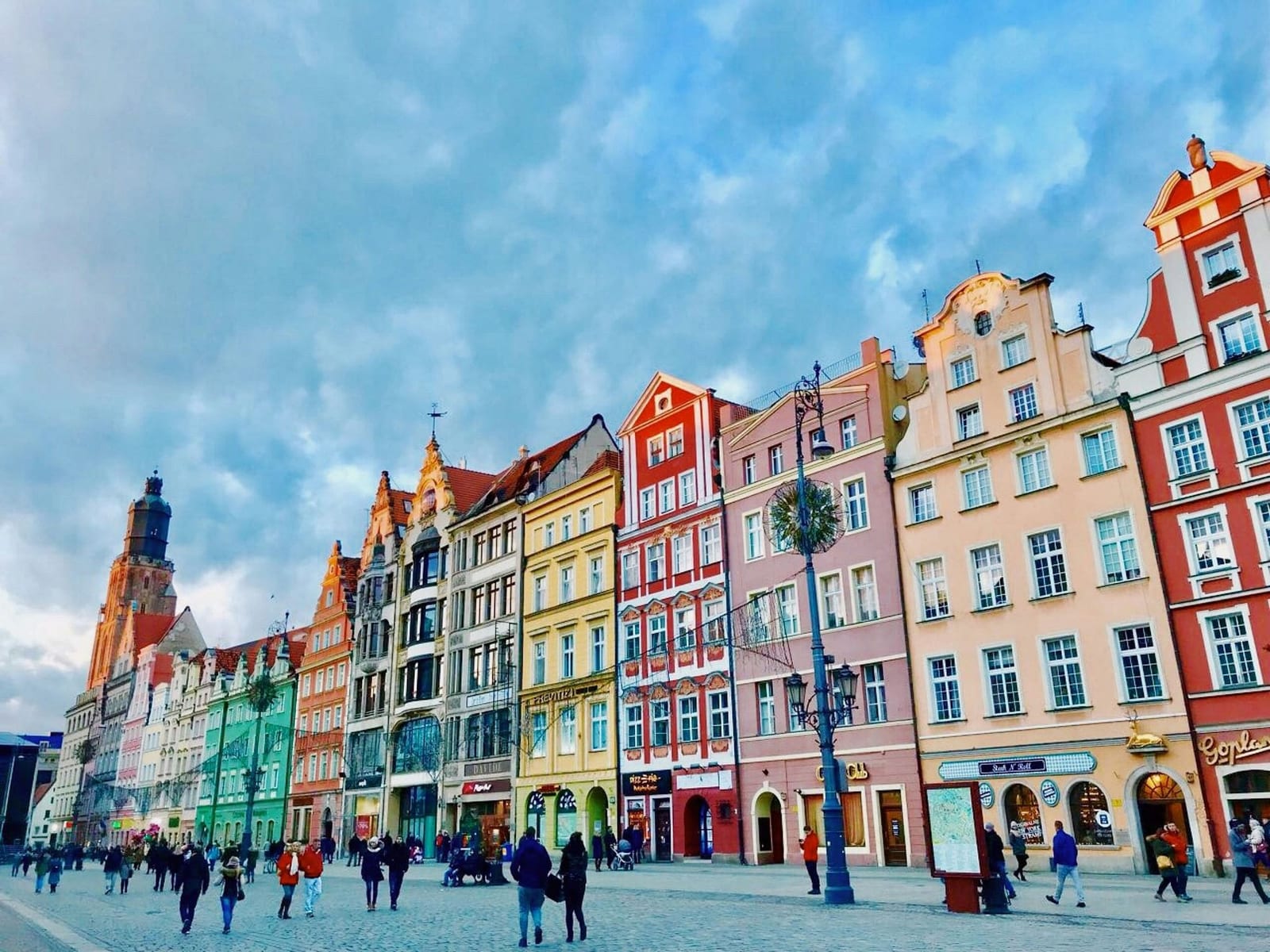 Wrocław's Market Square (Rynek), Poland, lined with vibrant Renaissance, Baroque, and Neoclassical kamienice townhouses in terracotta, mustard yellow, mint green, and pale blue, with ornate wrought-iron street lamps, cobblestone pavement, and the dark Gothic spire of St. Elizabeth's Church rising above the rooftops under a bright blue sky — book a fee-free Poland vacation rental on Houfy | Houfy — No Fees, Book Direct