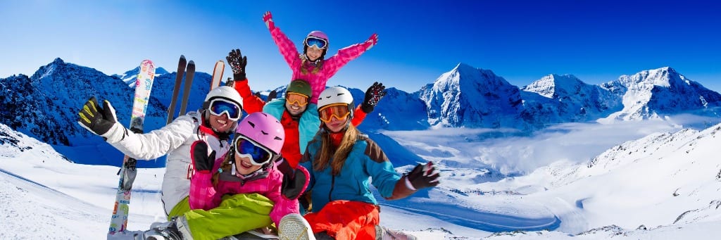 A joyful family of five in colorful ski gear cheering on a sun-drenched snowy mountain summit, with a panoramic backdrop of jagged alpine peaks and a brilliant cobalt blue sky — book a fee-free rental by owner on Houfy | Houfy — No Fees, Book Direct