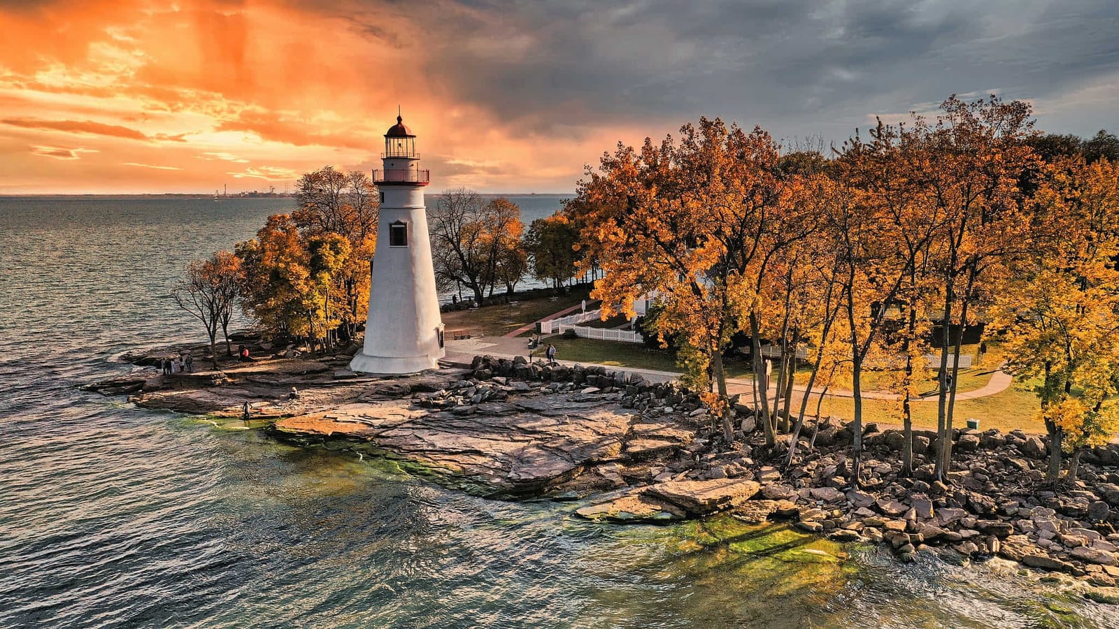 Point Judith Lighthouse and historic Coast Guard station at dusk in Narragansett, Rhode Island, with the octagonal granite and brownstone tower beacon glowing warm yellow, a stately white three-story station building with a bold fire-engine red gabled roof and American flag, tawny coastal beach grass in the foreground, and a dramatic charcoal and soft purple overcast sky over the calm Atlantic Ocean — book a fee-free Rhode Island vacation rental on Houfy | Houfy — No Fees, Book Direct