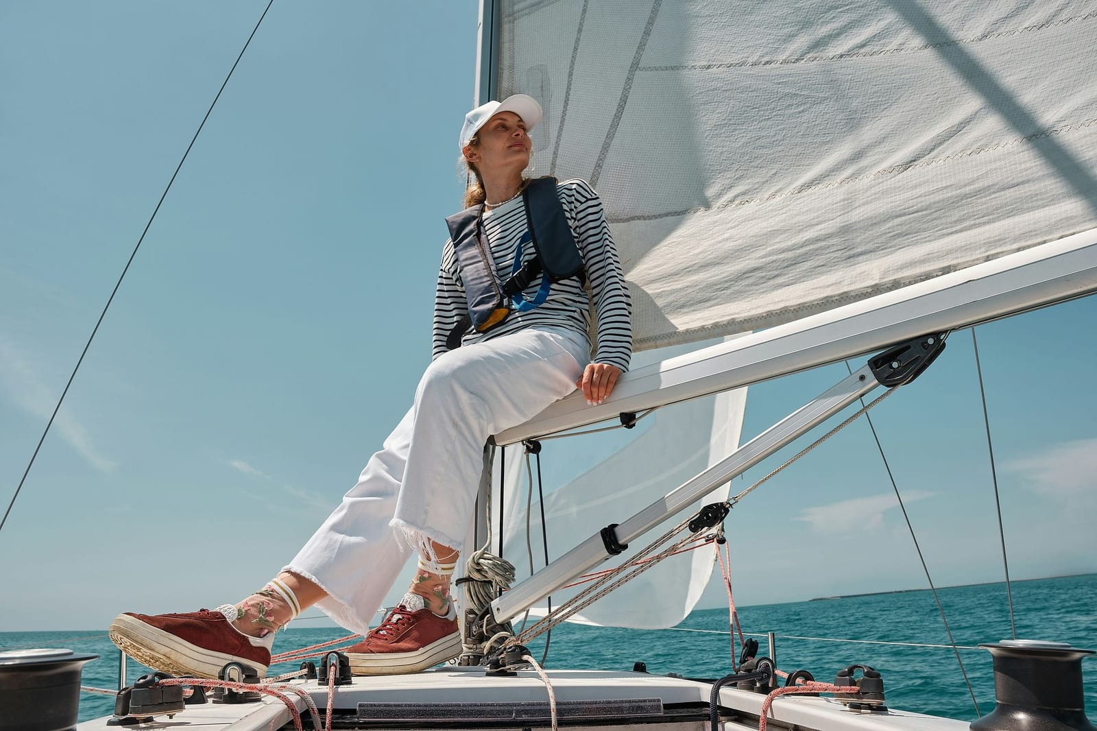 A woman in nautical-stripe shirt, navy life vest, and white cap perched on the boom of a modern sailboat with a billowing white mainsail over sparkling turquoise water under a clear blue sky — book a fee-free sailing vacation rental on Houfy | Houfy — No Fees, Book Direct