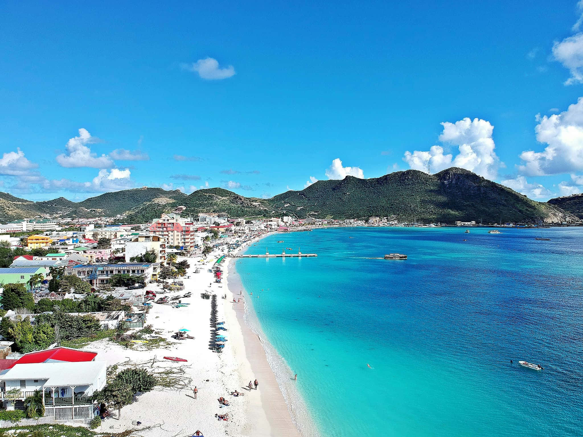 Aerial view of Great Bay in Philipsburg, Sint Maarten, with a wide crescent of white sand beach dotted with blue umbrellas and sunbathers, a long wooden pier extending into the bay, a brilliant turquoise-to-sapphire Caribbean water gradient, colorful terracotta and green rooftops lining the waterfront town, and lush emerald volcanic hills rising steeply behind the coast under a vivid blue sky — book a fee-free Sint Maarten vacation rental on Houfy | Houfy — No Fees, Book Direct