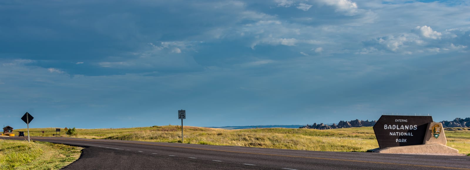 Badlands National Park sign greets travelers along a winding road under expansive cloudy skies, distinctive prairie grasses stretch towards the horizon, capturing the rugged beauty of this South Dakota landscape. | Houfy — No Fees, Book Direct