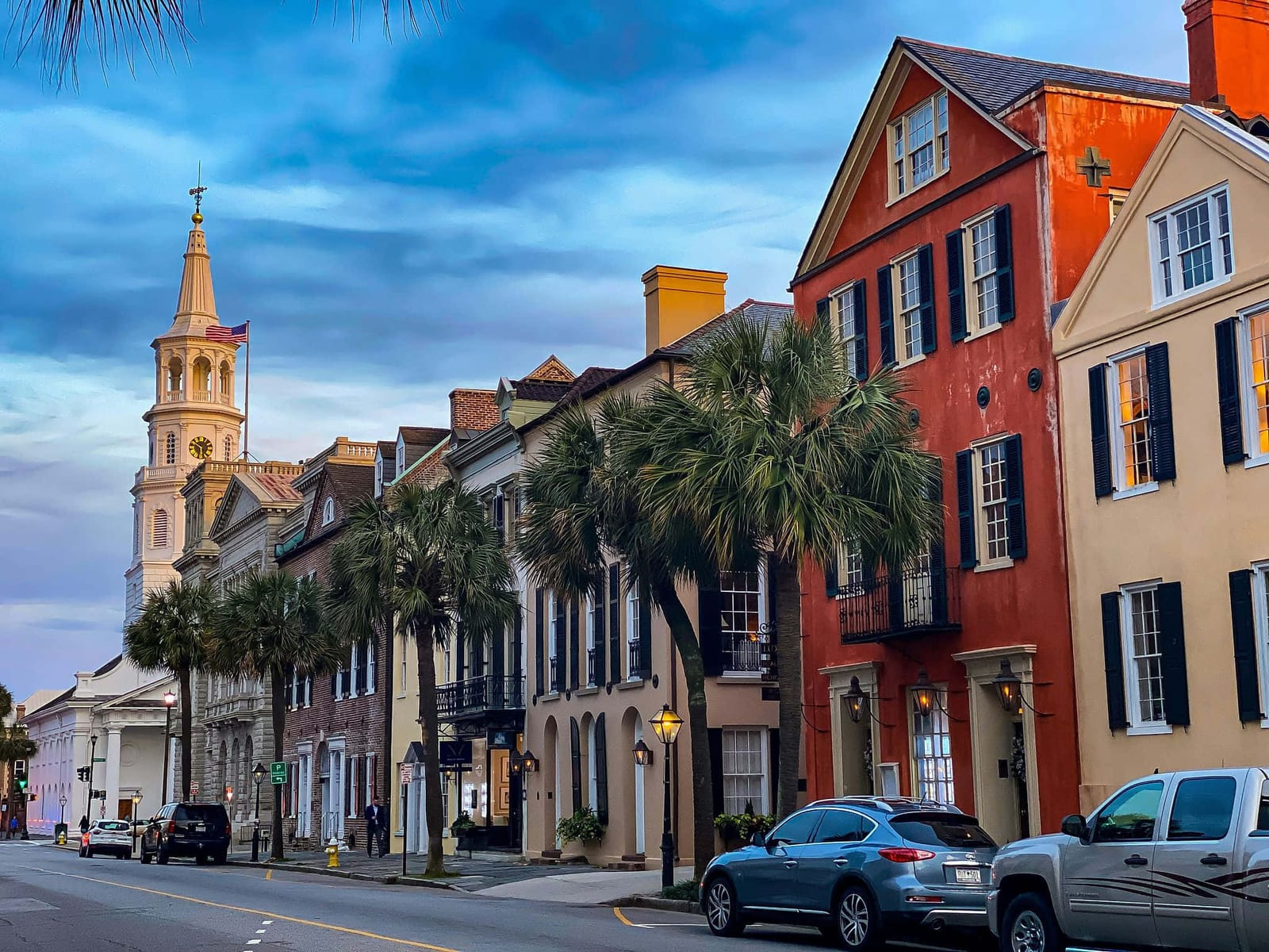Blue hour view down East Bay Street in historic Charleston, South Carolina, with a striking terracotta-red colonial building with dark green shutters and iron balconies beside pale yellow stucco facades, mature Sabal Palmetto state trees lining the sidewalk, warm golden gas lamp light, and the glowing white neoclassical steeple of St. Michael's Episcopal Church rising against a deep cerulean sky — book a fee-free South Carolina vacation rental on Houfy | Houfy — No Fees, Book Direct