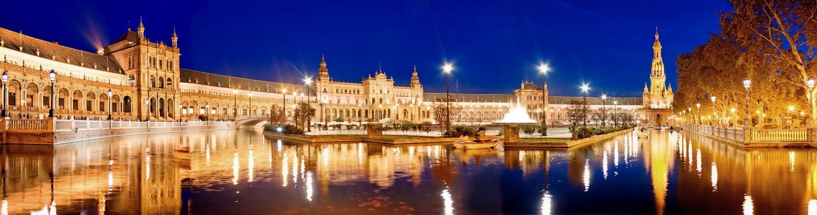 Panoramic blue hour view of Plaza de España in Seville, Spain, with golden-lit Renaissance and Moorish Revival architecture perfectly reflected in the calm canal below — book a fee-free Spain vacation rental on Houfy | Houfy — No Fees, Book Direct