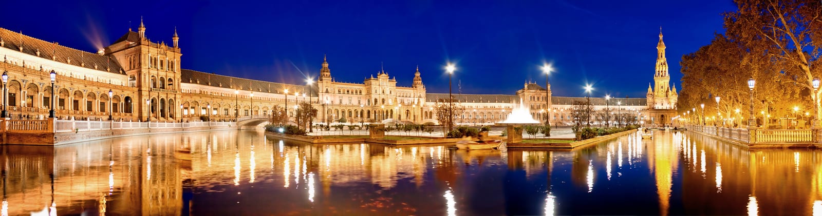 Panoramic blue hour view of Plaza de España in Seville, Spain, with golden-lit Renaissance and Moorish Revival architecture perfectly reflected in the calm canal below — book a fee-free Spain vacation rental on Houfy | Houfy — No Fees, Book Direct