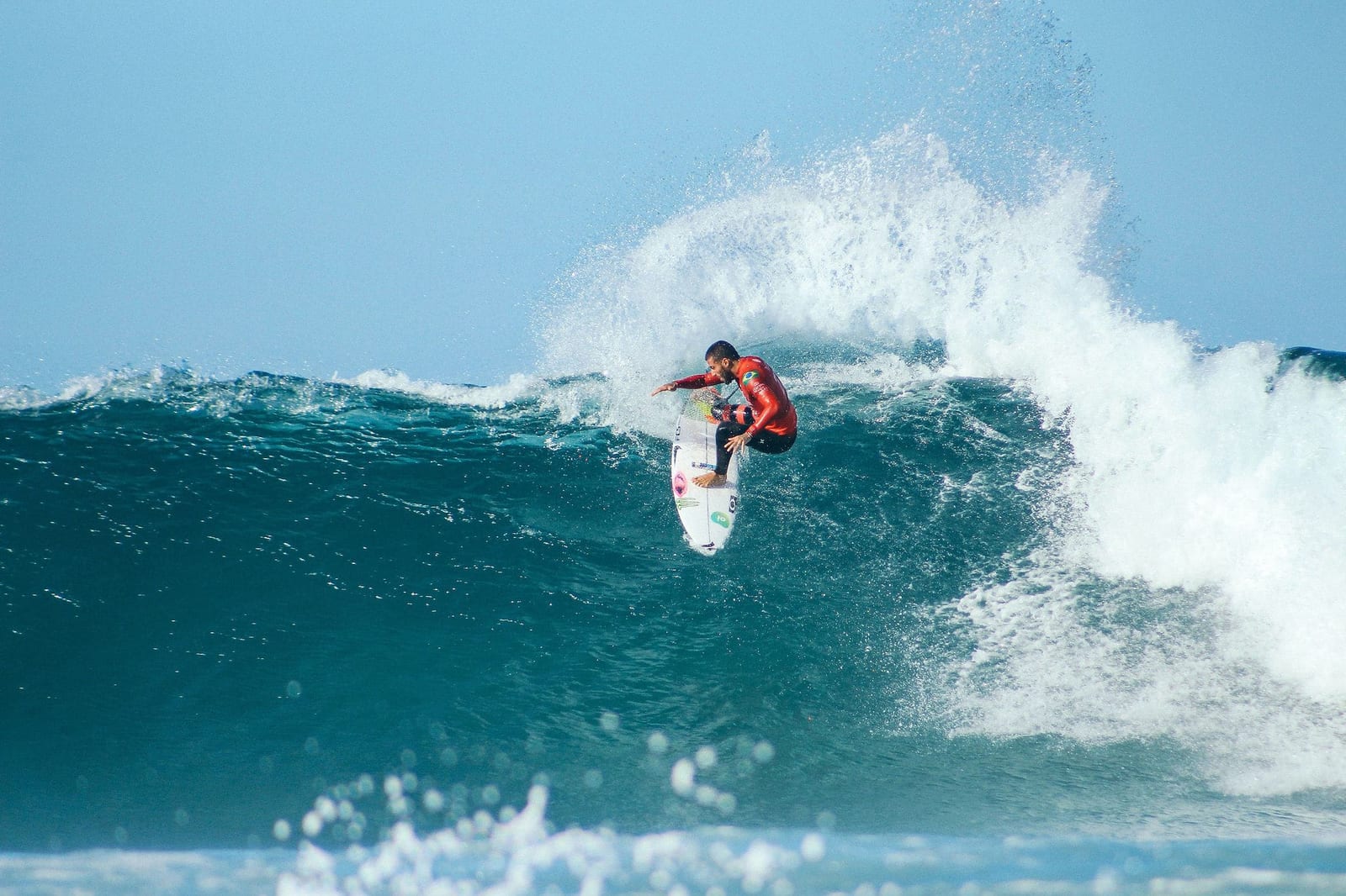 Male surfer in a red and black wetsuit launching an aerial off the exploding white lip of a massive turquoise wave, with a white shortboard tilted skyward and sea spray frozen mid-air — book a fee-free surfing vacation rental on Houfy | Houfy — No Fees, Book Direct