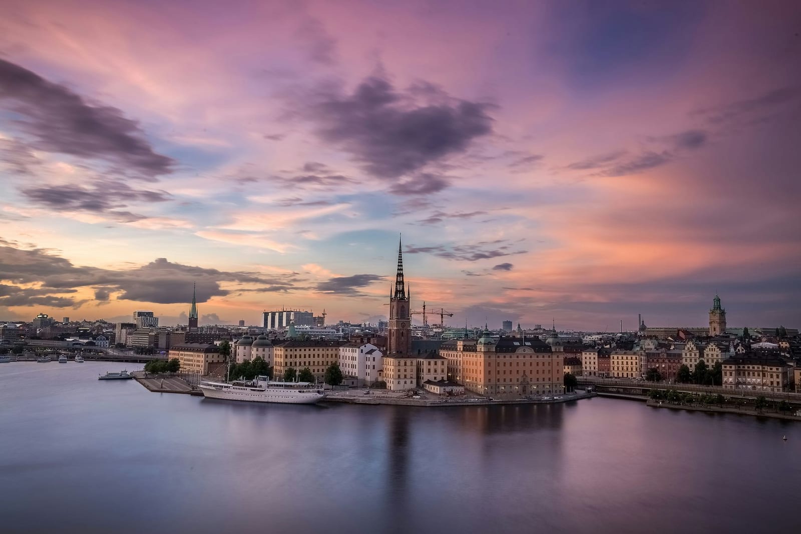 Twilight panorama of Stockholm, Sweden, with the dark latticed spire of Riddarholmen Church rising above ochre and terracotta historic buildings on Gamla Stan island, a vintage white passenger ship docked along the quay, and a long-exposure silk-smooth reflection of purple, pink, and amber sunset sky in the still waters of Lake Mälaren — book a fee-free Sweden vacation rental on Houfy | Houfy — No Fees, Book Direct
