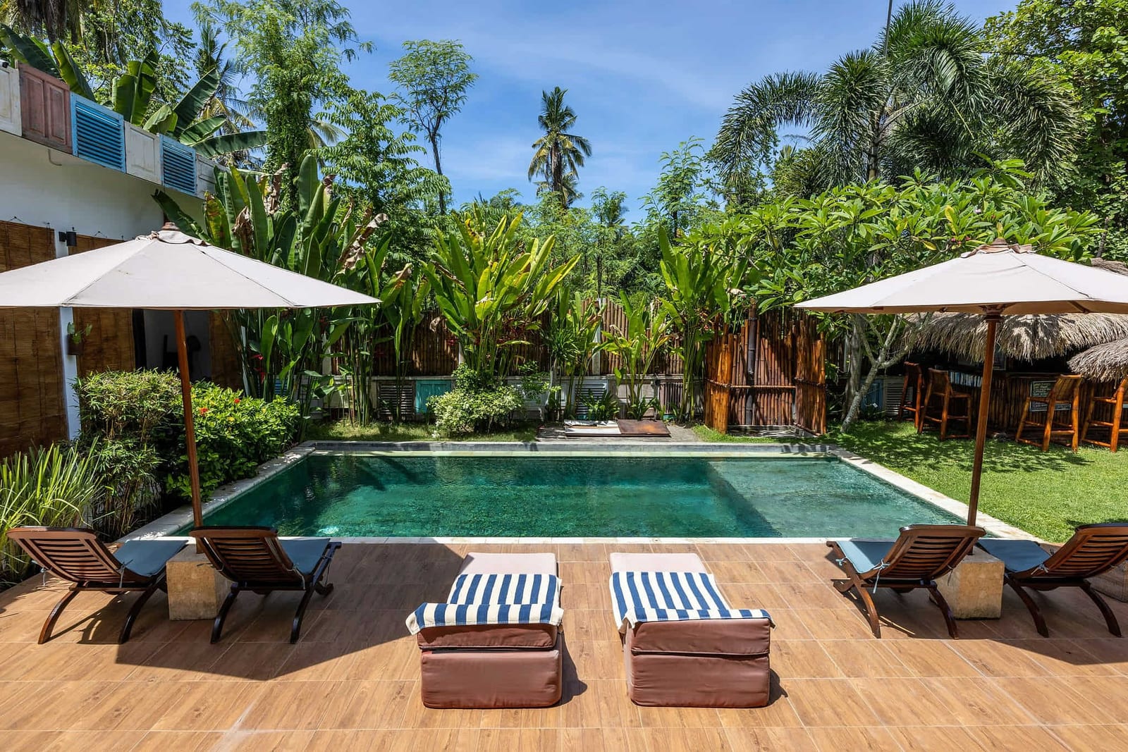 Pristine private swimming pool with rich emerald turquoise water, wooden sun loungers with blue cushions, white patio umbrellas, and a thatched tiki bar nestled in a lush tropical garden — book a fee-free swimming vacation rental on Houfy | Houfy — No Fees, Book Direct