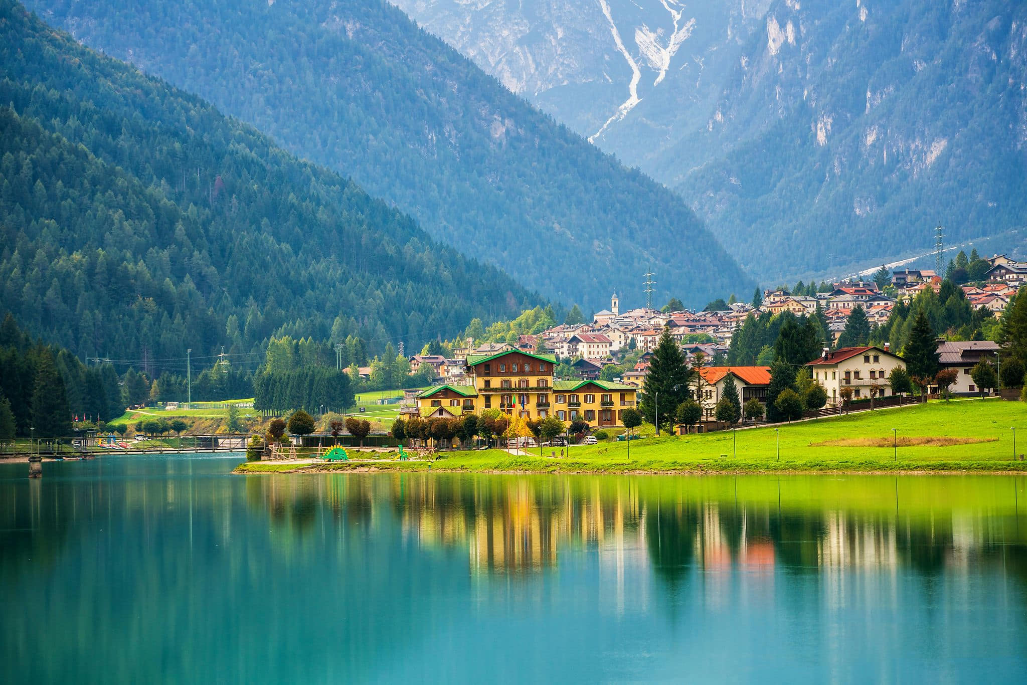 Serene Alpine village with traditional ochre-yellow chalets and a slender church steeple perfectly reflected in a glassy turquoise lake, framed by dense evergreen forests and misty blue-grey mountain peaks — book a fee-free Switzerland vacation rental on Houfy | Houfy — No Fees, Book Direct