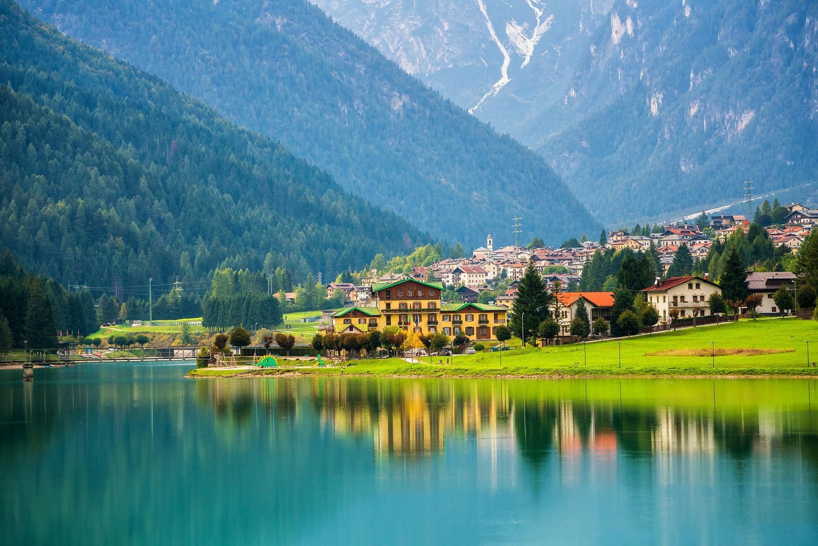 Serene Alpine village with traditional ochre-yellow chalets and a slender church steeple perfectly reflected in a glassy turquoise lake, framed by dense evergreen forests and misty blue-grey mountain peaks — book a fee-free Switzerland vacation rental on Houfy | Houfy — No Fees, Book Direct