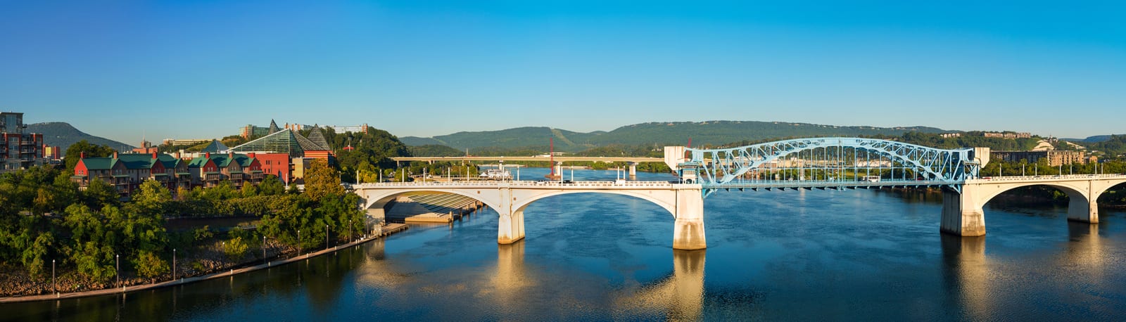 Panoramic view of Chattanooga, Tennessee, showcasing the Walnut Street Bridge spanning the Tennessee River, with lush greenery and Lookout Mountain in the background under a clear blue sky. | Houfy — No Fees, Book Direct