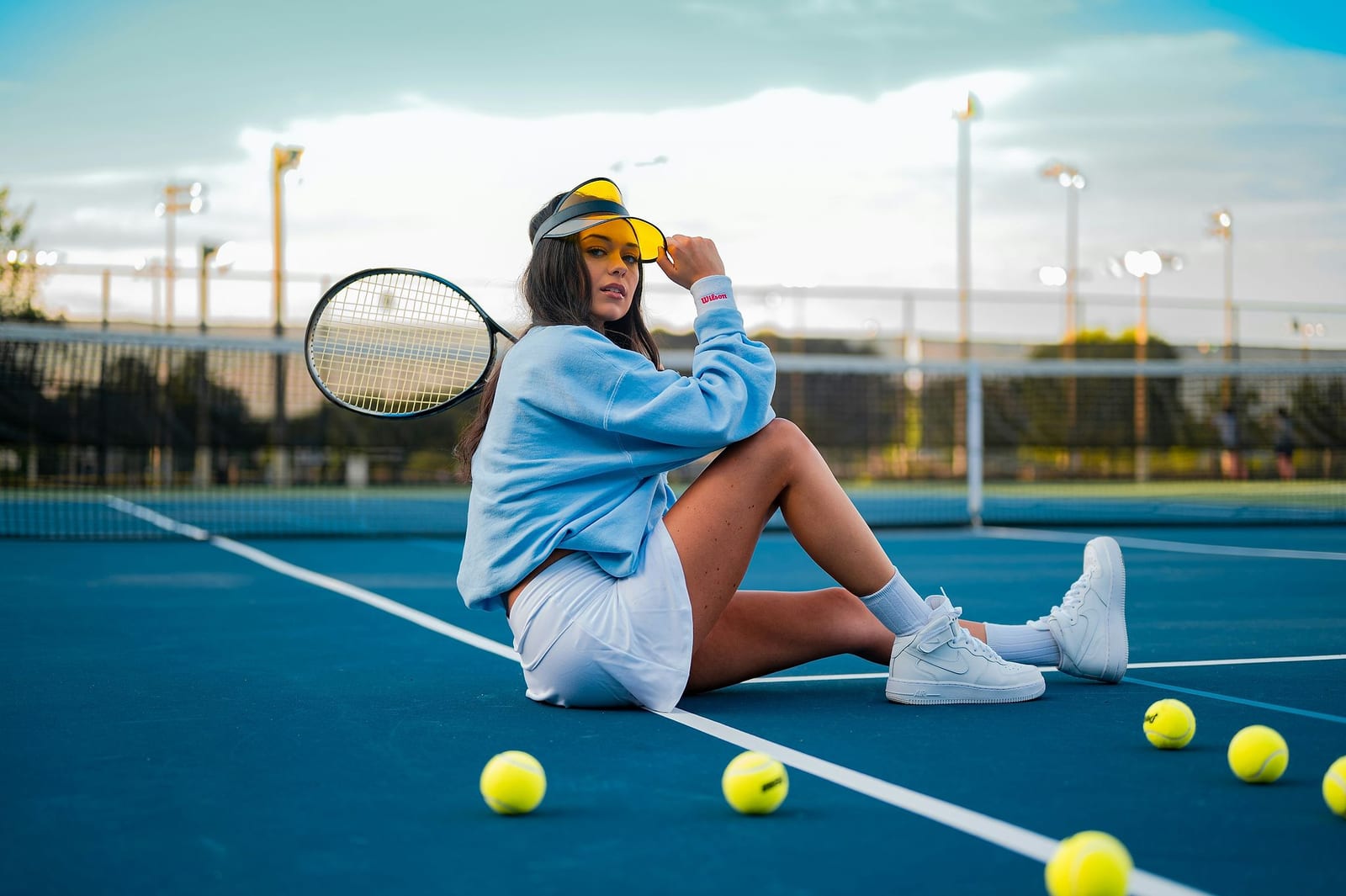 Stylish woman in a light blue sweatshirt and white tennis skirt sitting on a vibrant blue hardcourt at golden hour, black racket over her shoulder and neon yellow tennis balls scattered in the foreground — book a fee-free tennis vacation rental on Houfy | Houfy — No Fees, Book Direct