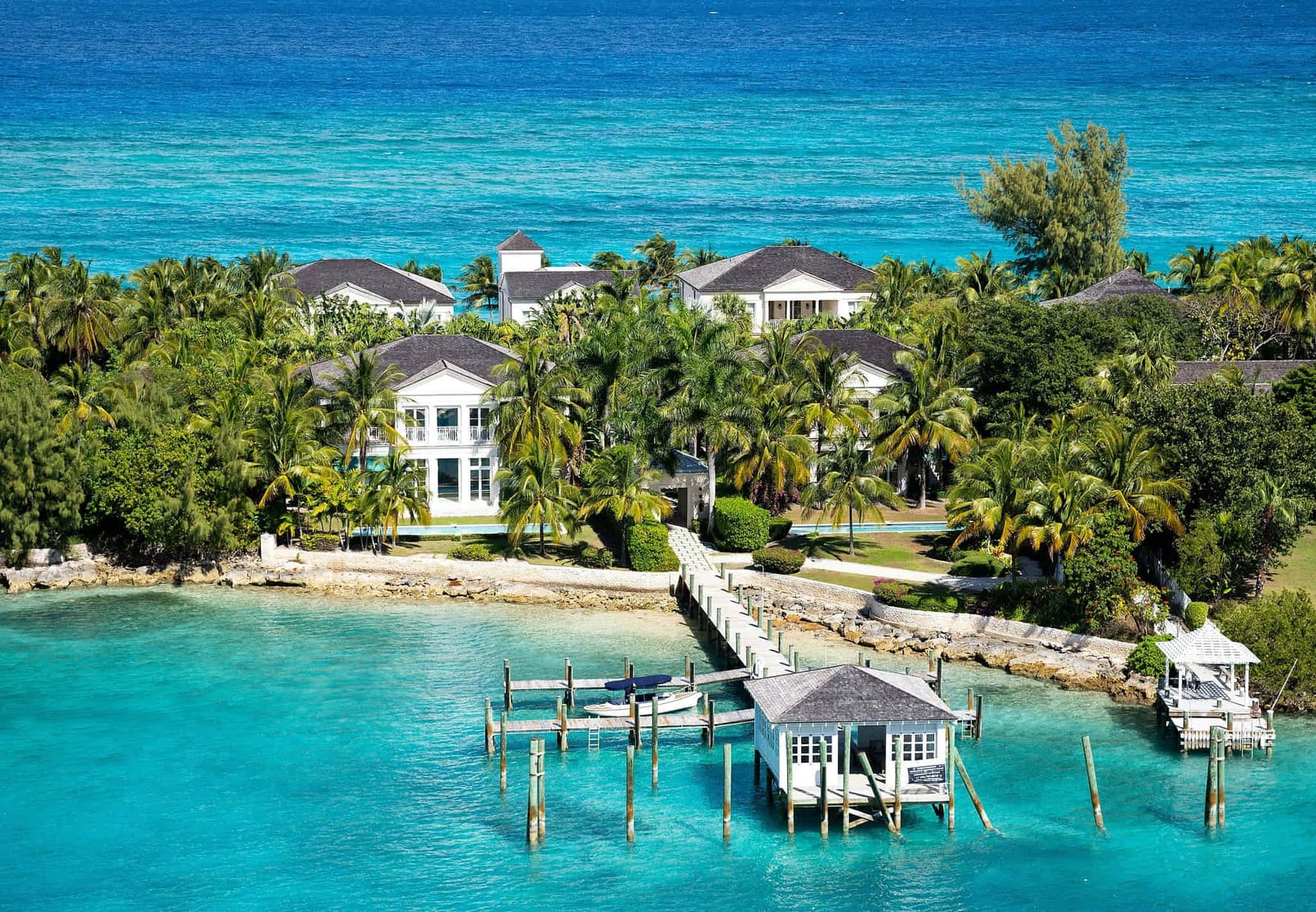 Aerial view of a secluded luxury Bahamian beachfront estate with elegant white two-story villas and dark shingled roofs nestled among towering palm trees and dense emerald tropical foliage, a weathered wooden pier extending over brilliant turquoise shallows to a white boathouse, and a dramatic gradient from translucent cyan to deep royal blue open ocean — book a fee-free Bahamas vacation rental on Houfy | Houfy — No Fees, Book Direct