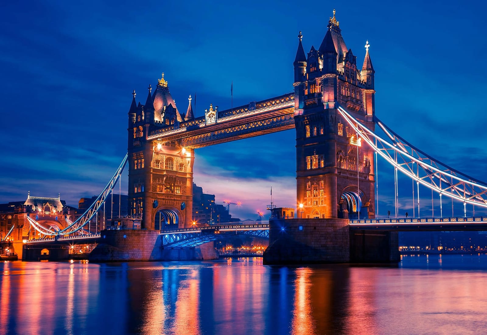 Blue hour long-exposure view of Tower Bridge, London, England, with Victorian Gothic stone towers glowing in warm golden light, illuminated suspension cables traced in white, and brilliant orange and cerulean blue reflections shimmering on the River Thames under a deep navy sky — book a fee-free United Kingdom vacation rental on Houfy | Houfy — No Fees, Book Direct