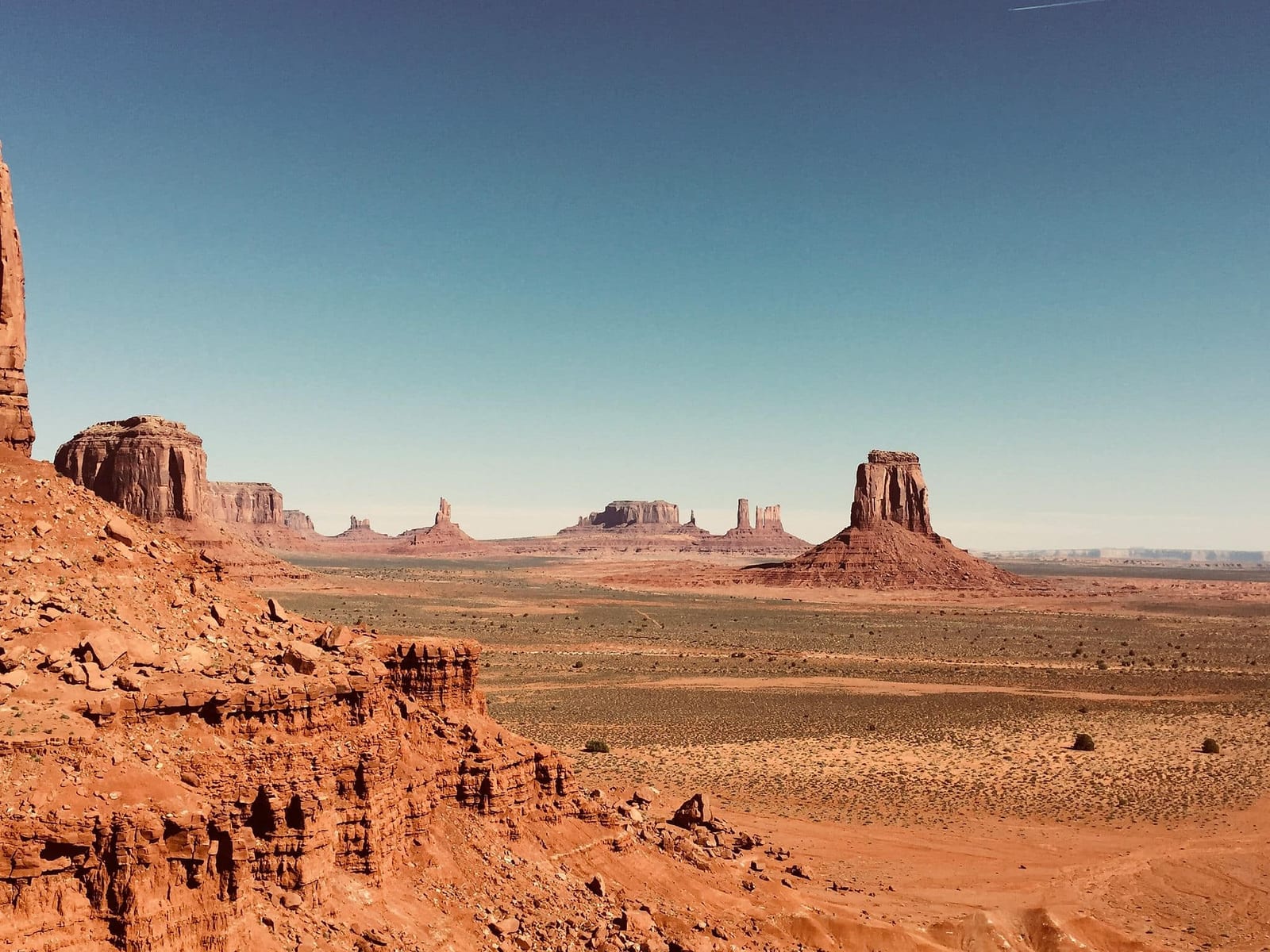 Panoramic view of Monument Valley Navajo Tribal Park on the Arizona-Utah border, with iconic rust-red sandstone buttes and mesas rising dramatically from a vast desert floor under a deep cerulean blue sky — book a fee-free United States vacation rental on Houfy | Houfy — No Fees, Book Direct
