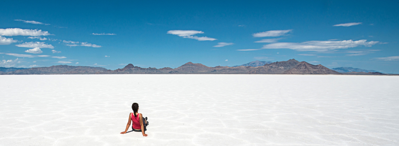 A lone traveler in a red shirt sits on the vast, white expanse of Salar de Uyuni under a clear blue sky, with distant mountains forming a dramatic backdrop in Bolivia's iconic salt flats. | Houfy — No Fees, Book Direct