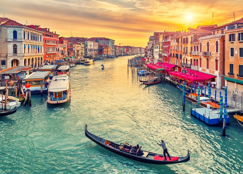 Gondola gliding through the vibrant Grand Canal in Venice at sunset, lined with historic colorful buildings and bustling waterside cafes, capturing the essence of Venetian charm and romance. | Houfy — No Fees, Book Direct