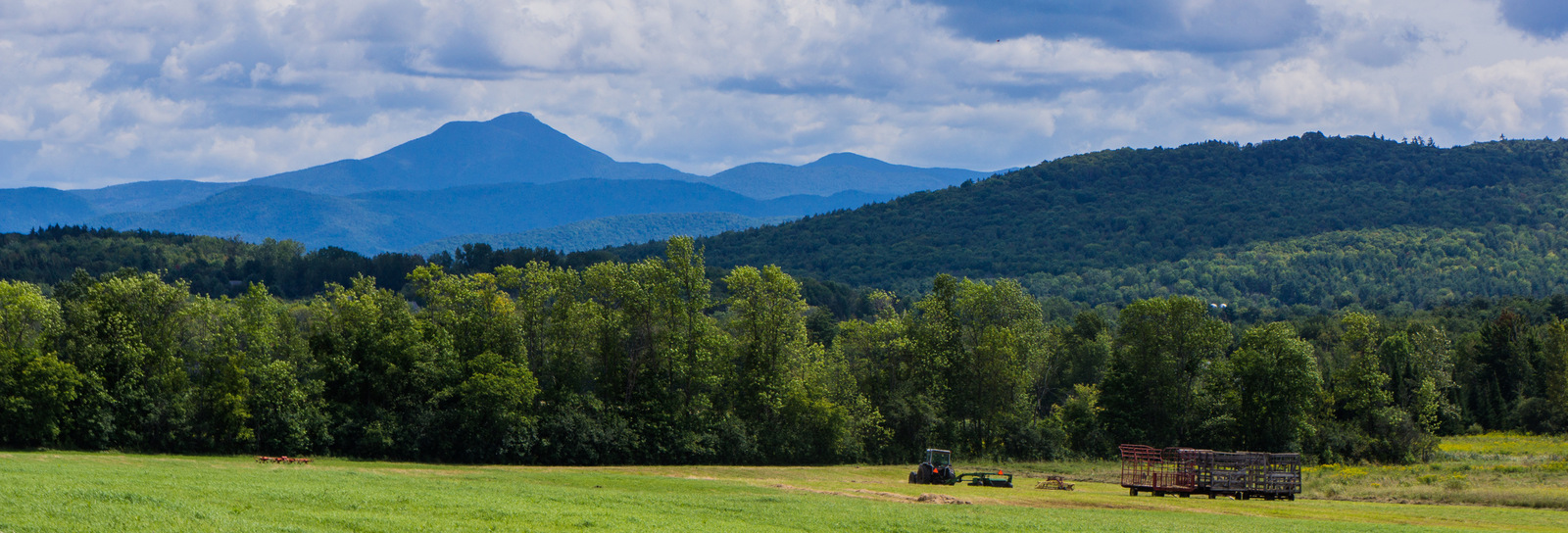 Vermont's picturesque landscape features lush green fields, dense forests, and distant mountain peaks framed by cloudy skies. A tractor and hay trailer add a rustic charm unique to this tranquil rural setting. | Houfy — No Fees, Book Direct
