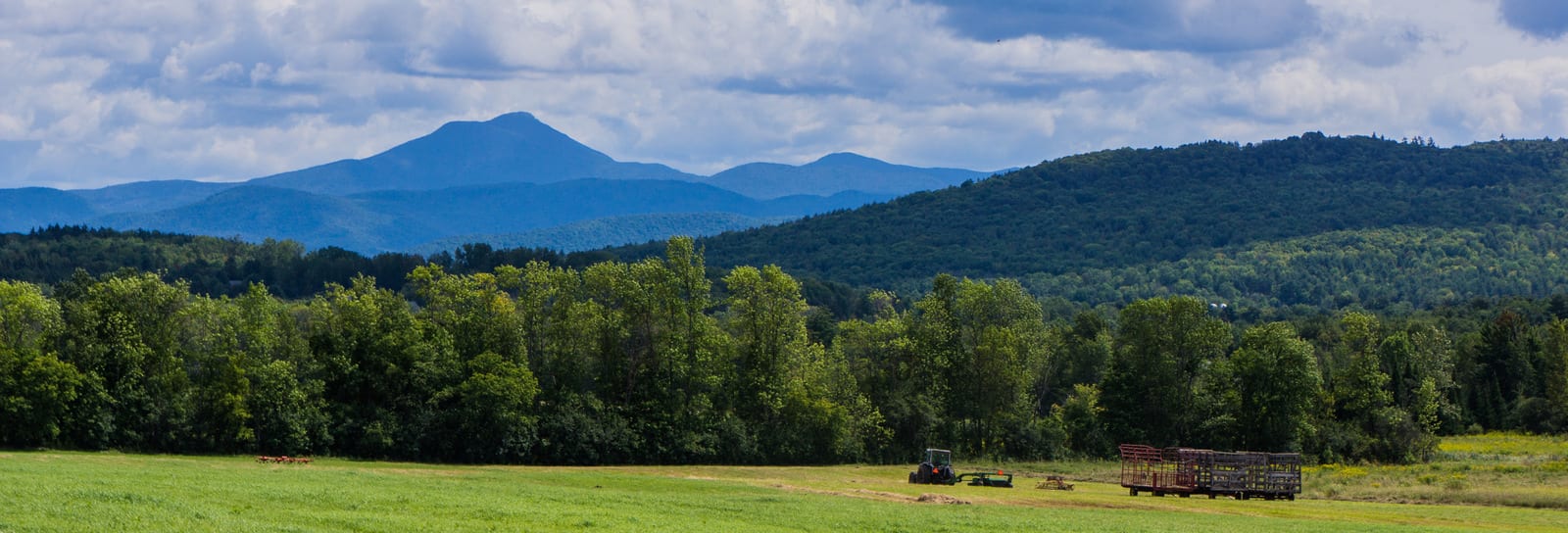 Vermont's picturesque landscape features lush green fields, dense forests, and distant mountain peaks framed by cloudy skies. A tractor and hay trailer add a rustic charm unique to this tranquil rural setting. | Houfy — No Fees, Book Direct