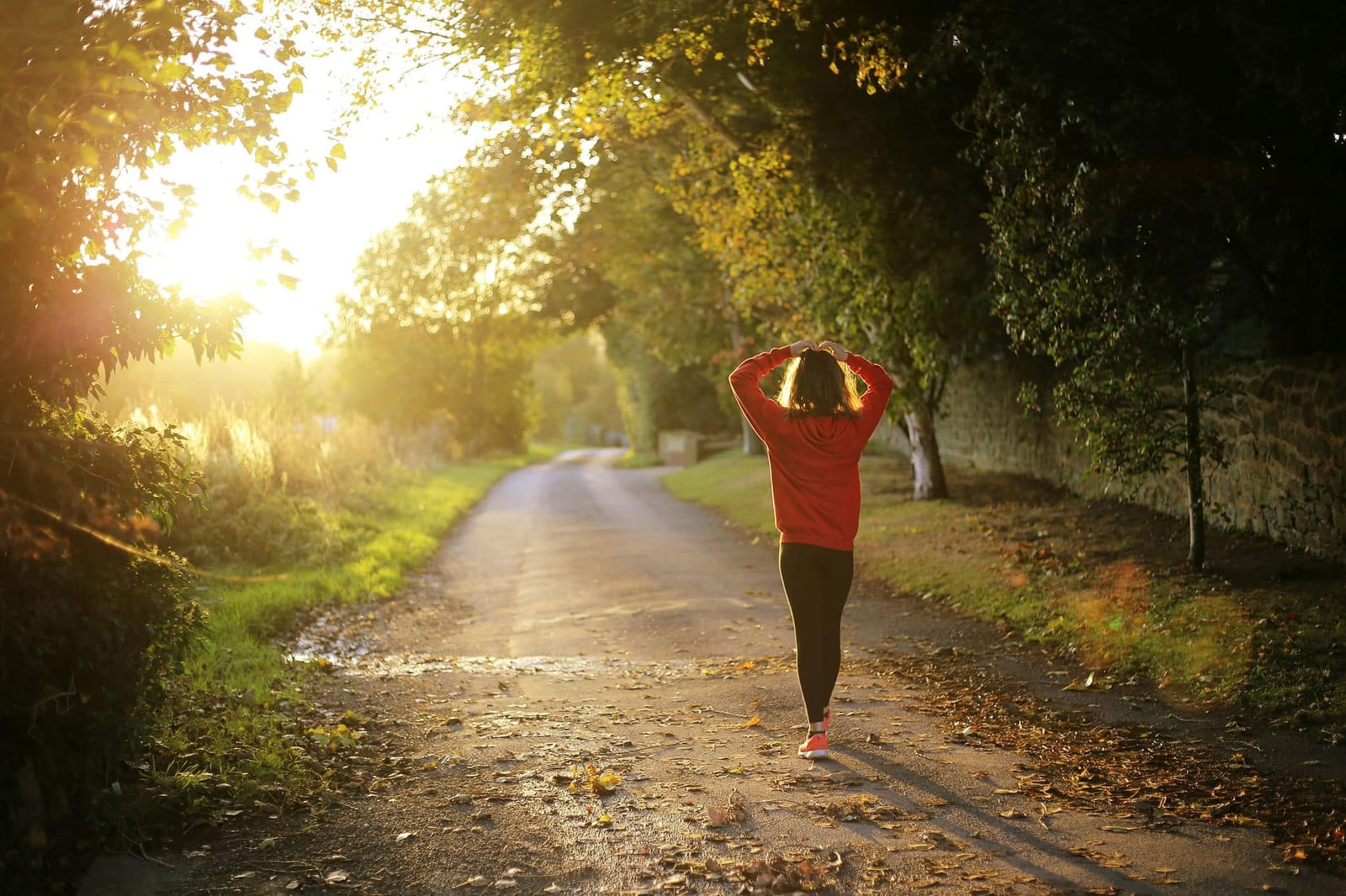 A woman in a vibrant red hoodie and black leggings walking down a sun-drenched narrow country lane at golden hour, flanked by a rustic stone wall, lush green trees, and golden lens flare light — book a fee-free walking vacation rental on Houfy | Houfy — No Fees, Book Direct