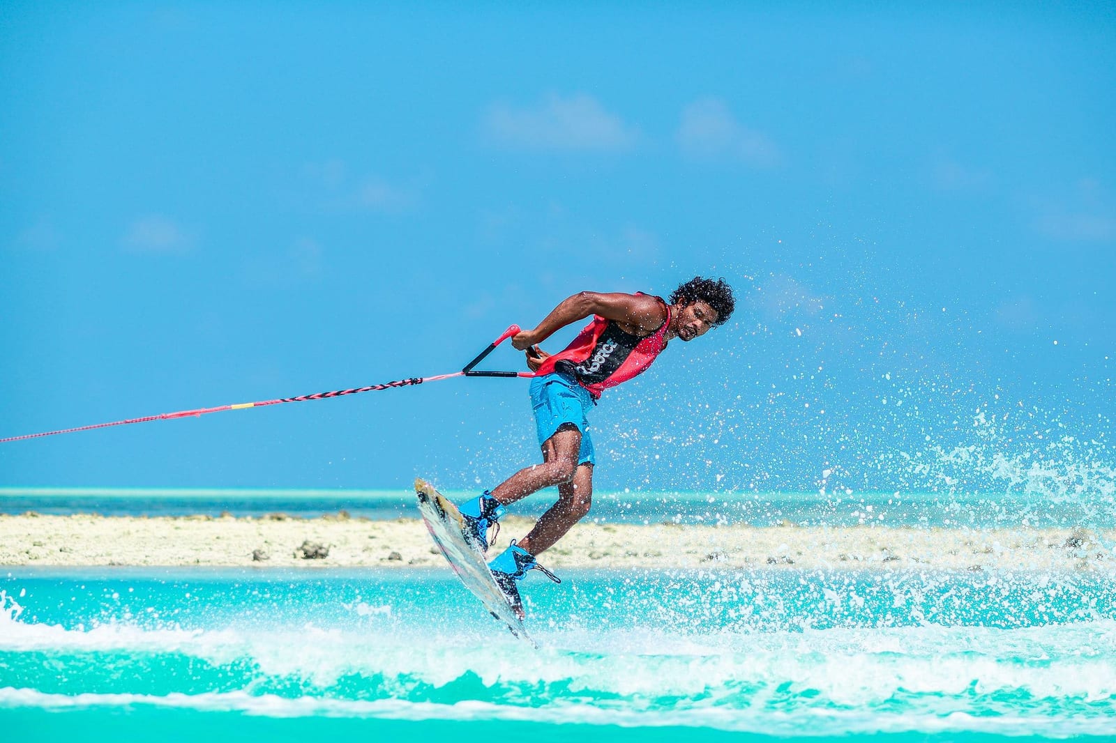 Athletic man in a red life vest and cyan board shorts wakeboarding at high speed across a brilliant turquoise tropical lagoon, with a massive white spray arc frozen mid-air and a white sand horizon in the distance — book a fee-free water skiing vacation rental on Houfy | Houfy — No Fees, Book Direct